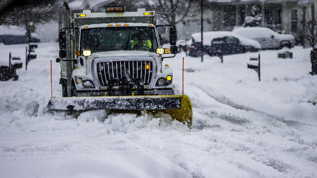 White snowplow service truck with orange lights and yellow plow blade clearing residential roads of snow while flakes are still falling 