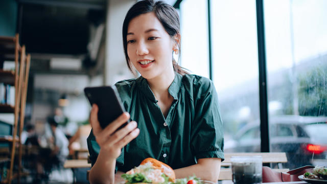 Woman managing online banking with mobile bank apps on smartphone while sitting in cafe having lunch 